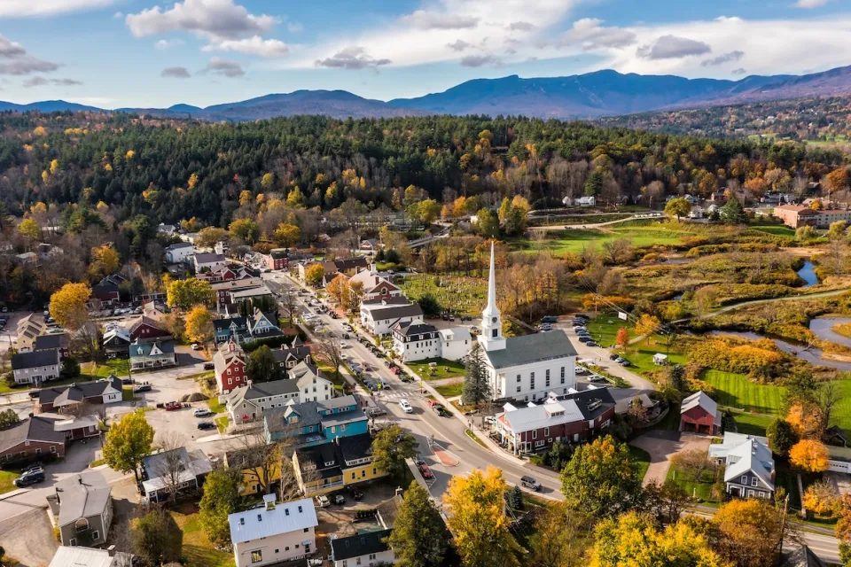 Aerial view of Stowe, Vermont, small rural town with tall church steeple and forest and mountains in background.